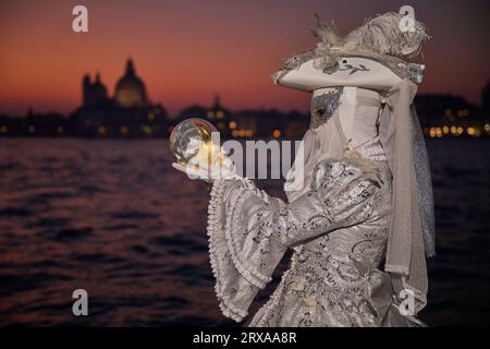 Genießen Sie venezianische Traditionen, während die Sonne über San Giorgio Maggiore untergeht, wo Masken und Magie sich im zauberhaften Karneval von 2023 vereinen. Stockfoto