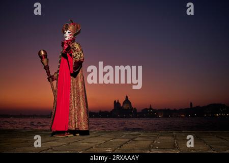 Genießen Sie venezianische Traditionen, während die Sonne über San Giorgio Maggiore untergeht, wo Masken und Magie sich im zauberhaften Karneval von 2023 vereinen. Stockfoto