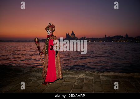 Genießen Sie venezianische Traditionen, während die Sonne über San Giorgio Maggiore untergeht, wo Masken und Magie sich im zauberhaften Karneval von 2023 vereinen. Stockfoto