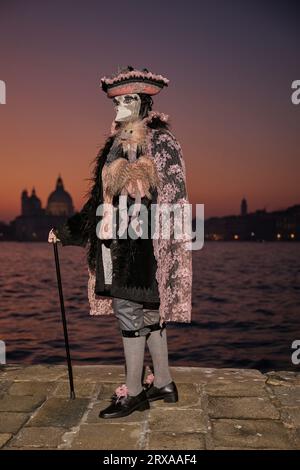 Genießen Sie venezianische Traditionen, während die Sonne über San Giorgio Maggiore untergeht, wo Masken und Magie sich im zauberhaften Karneval von 2023 vereinen. Stockfoto