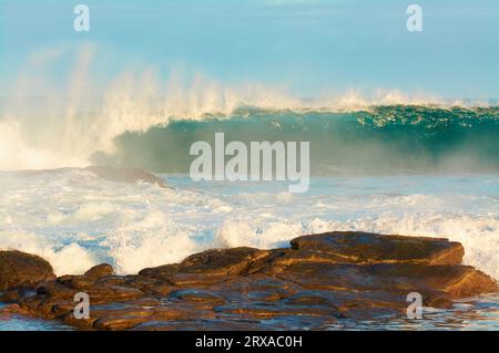 Eine große Welle bricht bei Sonnenaufgang mit Granitfelsen im Vordergrund, Prevelly Beach, Margaret River Region, Western Australia Stockfoto