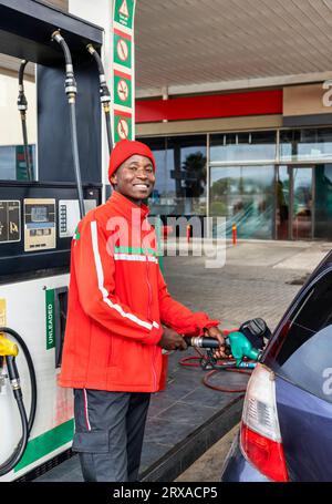 afroamerikaner in roter Uniform und ein lächelndes Gesicht an der Gaspumpe, das ein kleines Auto füllt Stockfoto