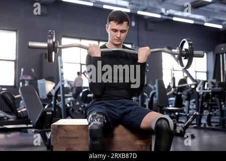 Der ehrgeizige Mann führt Langhantelübungen durch und sitzt auf einer Holzkiste. Übungen für das Training von Unterkörper und Kern. Kreuzschlitz Stockfoto