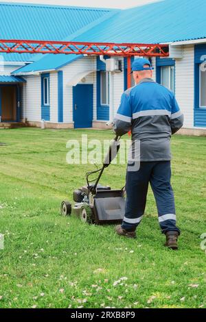 Arbeiter im Overall mäht Rasen mit Rasenmäher am Sommertag. Man mäht Gras auf dem Gebiet von Industrieanlagen. Stockfoto