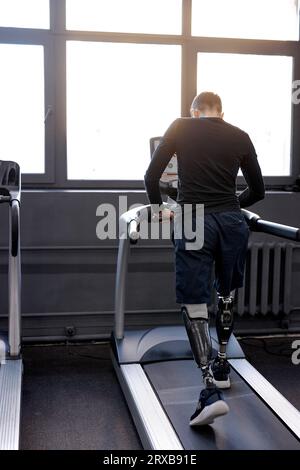 Muskulöser Mann in stilvoller Sportbekleidung, die auf Laufbändern im Fitnessstudio, Rückenansicht, Gesundheit und Körperpflege, in voller Länge, Sportler, die Rehabilitation ausüben Stockfoto
