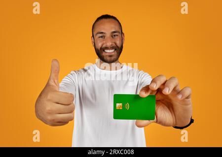 Glücklicher, gutaussehender junger europäer zeigt Kreditkarte, Daumen hoch, empfiehlt Finanzersparnisse Stockfoto