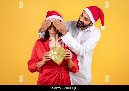 Glücklicher tausendjähriger arabischer Mann im Weihnachtsmannhut, der die Augen einer Frau mit Geschenk schließt, isoliert auf gelbem Studiohintergrund Stockfoto