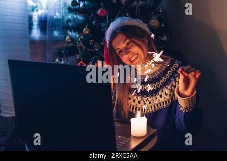 Feiern Sie Weihnachten und Neujahr online aus der Ferne mit einem Notebook. Eine junge Frau verbrennt zu Hause beim Telefonieren über das Internet an einem Weihnachtsbaum Stockfoto