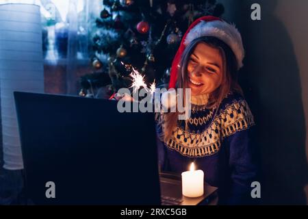 Feiern Sie Weihnachten und Neujahr online aus der Ferne mit einem Notebook. Eine junge Frau verbrennt zu Hause beim Telefonieren über das Internet an einem Weihnachtsbaum Stockfoto