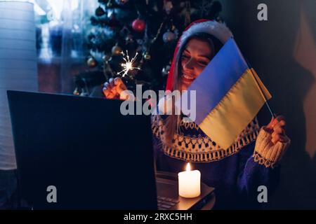 Feiern Sie Weihnachten und Neujahr online aus der Ferne mit einem Notebook. Eine Frau brennt zu Hause mit ukrainischer Flagge. Ich gratuliere der Familie Abroa Stockfoto