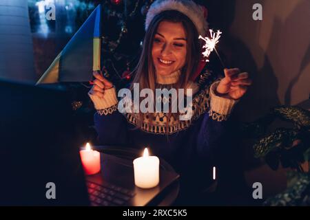 Wir feiern Weihnachten und Neujahr online aus der Ferne mit einem Laptop während des Krieges. Eine Frau brennt zu Hause mit ukrainischer Flagge. Ich rufe den Ehemann an Stockfoto