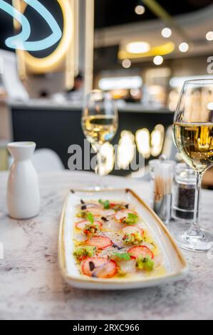 Wolfsbarsch-Ceviche mit Erdbeeren, Kiwi und Mangosalsa auf einem Marmortisch in einem Restaurant und einem Glas Weißwein Stockfoto