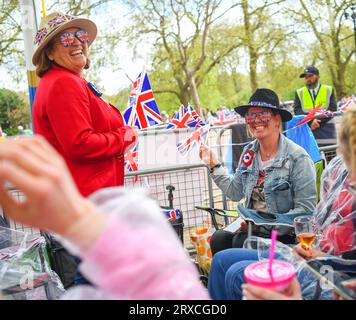 Zwei Frauen lächeln und feiern auf dem Campingplatz in der Mall London UK vor der Krönung von König Charles, gekleidet in Union Jack Brillen, die Fahnen schwenken Stockfoto