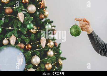 Frau, die den großen Weihnachtsbaum dekoriert. Weibchen mittleren Alters, das eine grüne Kugel in der Hand hält und am Baumzweig hängt. Leerzeichen kopieren. Grußkarte. Nahaufnahme Stockfoto