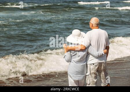 Ein älteres Paar steht am Strand am Meer, schaut auf die Wellen und entspannt sich. Rückansicht von hinten. Stockfoto