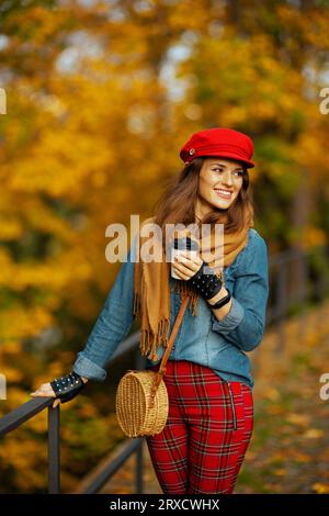 Hallo Herbst. Lächelnde, trendige Frau in Jeans-Hemd und roter Mütze mit Schal, Handschuhen, Tasche und Kaffee im Stadtpark. Stockfoto