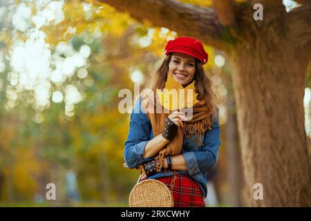 Hallo Herbst. Porträt einer glücklichen modernen Frau in Jeans-Hemd und roter Mütze mit Herbstblatt, Schal, Handschuhen und Tasche in der Nähe von Baum im Stadtpark. Stockfoto