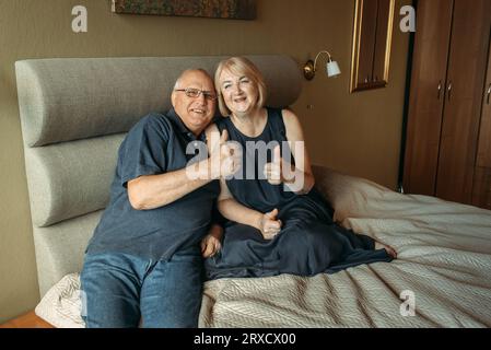 Ein älteres Paar sitzt auf einem Bett und zeigt eine Geste mit den Händen - Daumen hoch, wie. Stockfoto