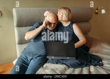 Glückliches Paar älterer Rentner, die online einkaufen und gemeinsam mit einem Laptop Artikel auswählen. Der Mann passt seine Brille an. Stockfoto