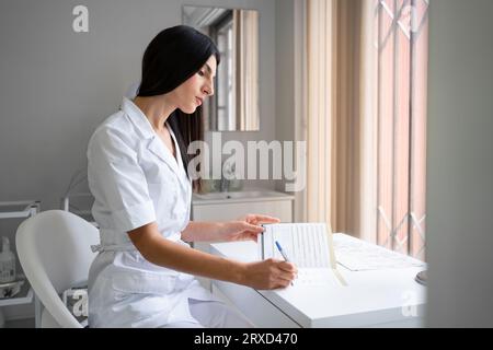 Ärztin füllt Patientenakte auf. Junge brünette Therapeutin in weißer Uniform, die neben dem Fenster sitzt. Kosmetologin, die Patienteninformationen aufschreibt, sitzt in der Arztpraxis Stockfoto