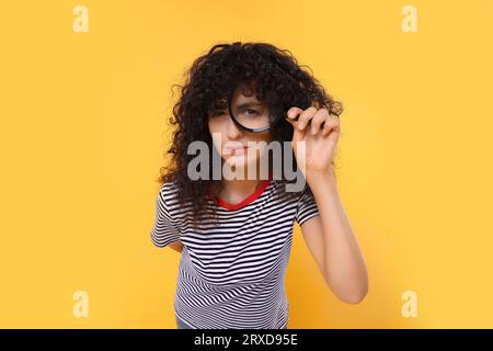 Neugierige junge Frau, die durch Lupenglas auf gelbem Hintergrund schaut Stockfoto