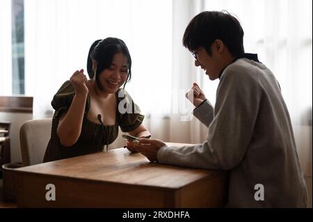 Zwei fröhliche junge asiatische Freundinnen und Freundinnen spielen gerne mobile Spiele oder schauen sich etwas Interessantes an, während sie sich in einem Café entspannen Stockfoto