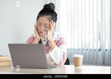 Eine müde und gestresste junge asiatische Frau leidet unter Kopfschmerzen und Augenschmerzen, nachdem sie stundenlang auf ihren Laptop-Bildschirm starrt und sich überfordert fühlt. Stockfoto