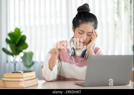 Eine müde junge asiatische Frau leidet unter Kopfschmerzen und Augenschmerzen, nachdem sie stundenlang auf ihren Laptop-Bildschirm starrt, sich überfordert und erschöpft fühlt. Stockfoto