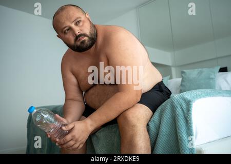 Müder, fetter Mann sitzt nach dem Training zur Gewichtsreduktion mit Wasserflasche auf dem Bett und denkt über das Ziel der Zukunft nach Stockfoto