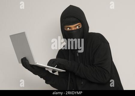 Porträt eines mysteriösen Mannes, der einen schwarzen Hoodie und eine Maske trägt, der auf einem Laptop hackt und einen Computer hält. Cyber-Sicherheitskonzept. Stockfoto