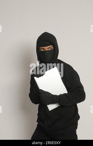 Mysteriöser Räuber-Dieb-Mann mit schwarzem Hoodie und Maske, der Laptop klaut und sich rausschleicht. Isoliertes Bild auf grauem Hintergrund Stockfoto