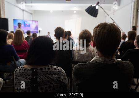 Hintergrund nicht erkennbarer Personen, die mit dem Rücken zur Kamera stehen und der Bühne zugewandt sind, auf der die Lautsprecher sprechen. Geschäftsereignis. Hochwertige Fotos Stockfoto