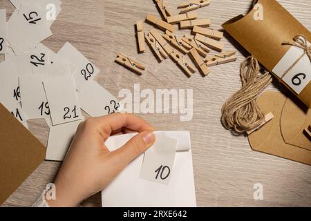 Eine nicht wiedererkennbare junge Frau klebt die Nummer auf die Handwerkstasche, wird mit einer Wäscheklammer befestigt. Weiblich macht Kraftpapier für hausgemachte Adventskalender mit eigenen Händen Schritt für Schritt selbst gemacht. Vorbereitung auf das weihnachtskonzept. Saisonale Aktivitäten für Kinder, Familienurlaub im Winter. Umweltfreundlich Geschenke. Öffnen Sie das Paket jeden Tag Stockfoto