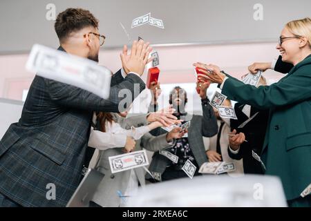 Fröhlicher afrikanisch-amerikanischer männlicher Boss in Sonnenbrille und Anzug, der mit Geldkanonen schießt. Fröhliches Business-Team aus verschiedenen Kollegen Stockfoto