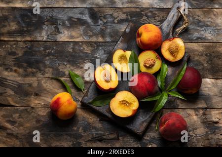 Frisch reife dunkelrote Pfirsiche mit Blättern auf Schneidebrett und Holztisch. Ganze Früchte und Hälften saftiger, heller orangener, köstlicher Pfirsiche auf dem Eingang Stockfoto