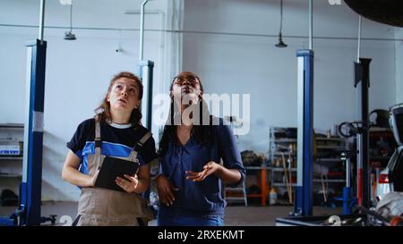 Erfahrener Mechaniker und Kunde, der unter dem Fahrzeug in der Garage steht und mit dem Laptop neue Teile bestellt, nachdem er Probleme gefunden hat. Experten helfen Kunden bei der Suche nach Komponenten online, Dolly in Shot Stockfoto