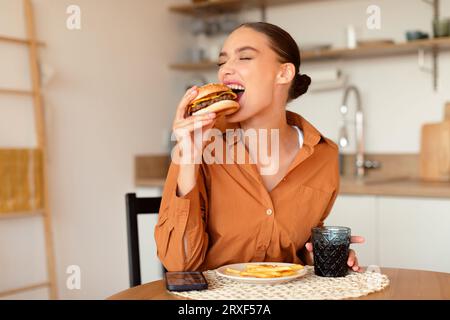 Junk Food. Dame, die leckeren Burger isst, ungesundes Fastfood hält und beißt, am Tisch in der Küche sitzt, freien Platz Stockfoto