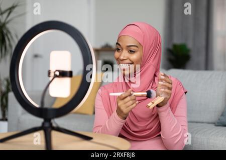 afroamerikanische muslimische Make-up-Künstlerin, die Videos für ihren Beauty-Blog auf dem Smartphone aufnimmt und ein neues Beauty-Produkt testet Stockfoto