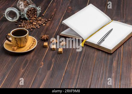 Notebook mit Stift öffnen. Tasse Kaffee, Kaffeebohnen in einem Glas. Auf dem Tisch gewürzt Sternanis und Stücke braunen Zuckers. Dunkler hölzerner Hintergrund. Co Stockfoto