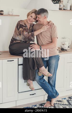 Familie genießt zarten Moment, schöne glückliche Frau mittleren Alters umarmt grauhaarigen Mann sitzend auf Küchentisch Stockfoto