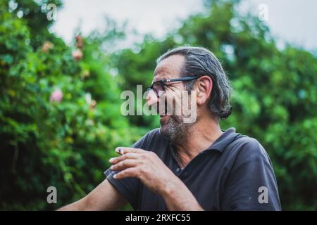 Ein älterer Gentleman im Alter von 60 Jahren mit Brille und langem Haar genießt die Natur inmitten von üppigen grünen Bäumen und strahlt ein freudiges Lächeln aus Stockfoto