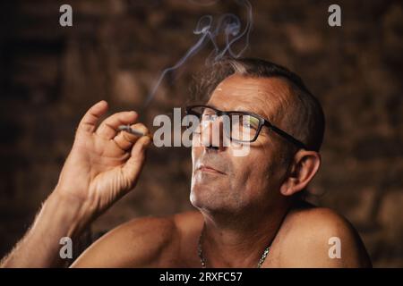 Ein älterer Mann, etwa 60 Jahre alt, serbischer Nationalität mit langen Haaren und Brille, raucht nachdenklich eine Zigarette auf dunklem Hintergrund. Künstler, cre Stockfoto