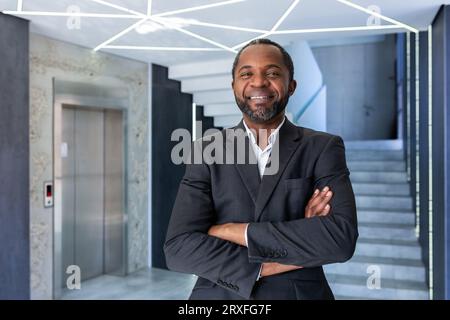 Porträt eines Reifen und erfolgreichen Geschäftsmannes im Büro, Chef lächelnd und mit gekreuzten Armen in die Kamera schauend, afroamerikanischer Mann im Business-Anzug glücklich über erfolgreiche Erfolge. Stockfoto