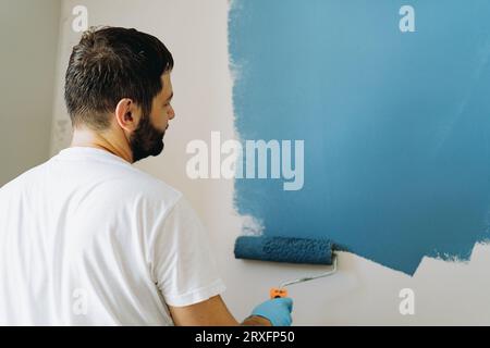 kaukasischer bärtiger Mann malte Wand mit Malerwalze. Malerwohnung, renovierendes Haus mit blauer Farbe. Bild mit selektivem Fokus Stockfoto