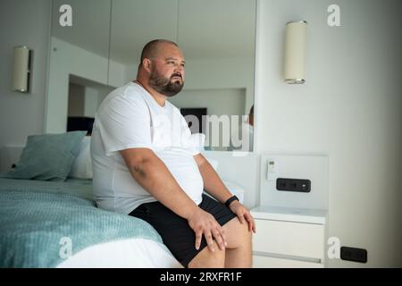 Fetter Mann, der auf dem Bett sitzt und ein unzufriedenes Gesicht hat. Der Typ wachte von Hitze auf, die bei hohen Temperaturen verstopfte Stockfoto