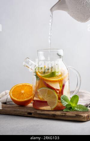 Hausgemachter, gesunder heißer Obsttee mit frischen, reifen Orangen, Apfeln, Minzblättern und Thymianzweigen in Glas-Teekanne oder Wasserkocher auf grauem Küchenhintergrund Stockfoto
