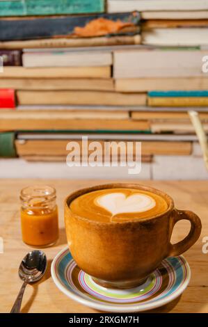 Rustikaler ockerfarbener Cappuccino-Becher mit Karamell und Himalaya-Salz, geformter Schaum auf Teller, Holz und Buchhintergrund - Taza de Capuchino rústica ocre con caram Stockfoto