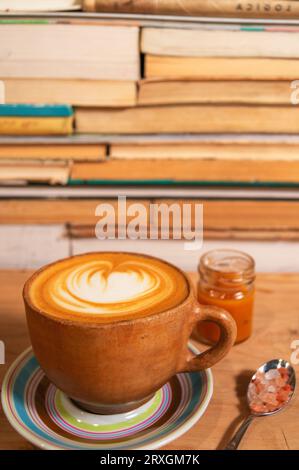 Rustikaler ockerfarbener Cappuccino-Becher mit Karamell und Himalaya-Salz, geformter Schaum auf Teller, Holz und Buchhintergrund - Taza de Capuchino rústica ocre con caram Stockfoto