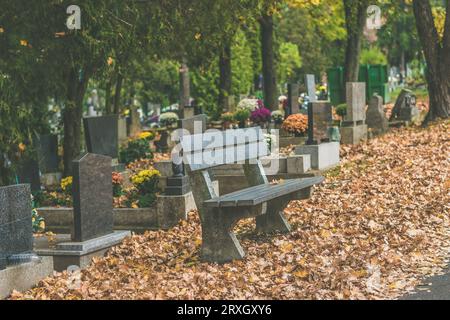 Leere Bank auf dem herbstlichen Friedhof mit bunten Dekorationen auf Gräbern und orangefarbenen Blättern auf dem Boden Stockfoto