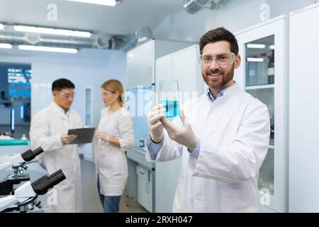Porträt eines jungen männlichen Chemikers, der in einem Labor steht und einen Kolben mit einer unbekannten blauen Substanz in den Händen hält und die Kamera lächelnd anschaut. Stockfoto